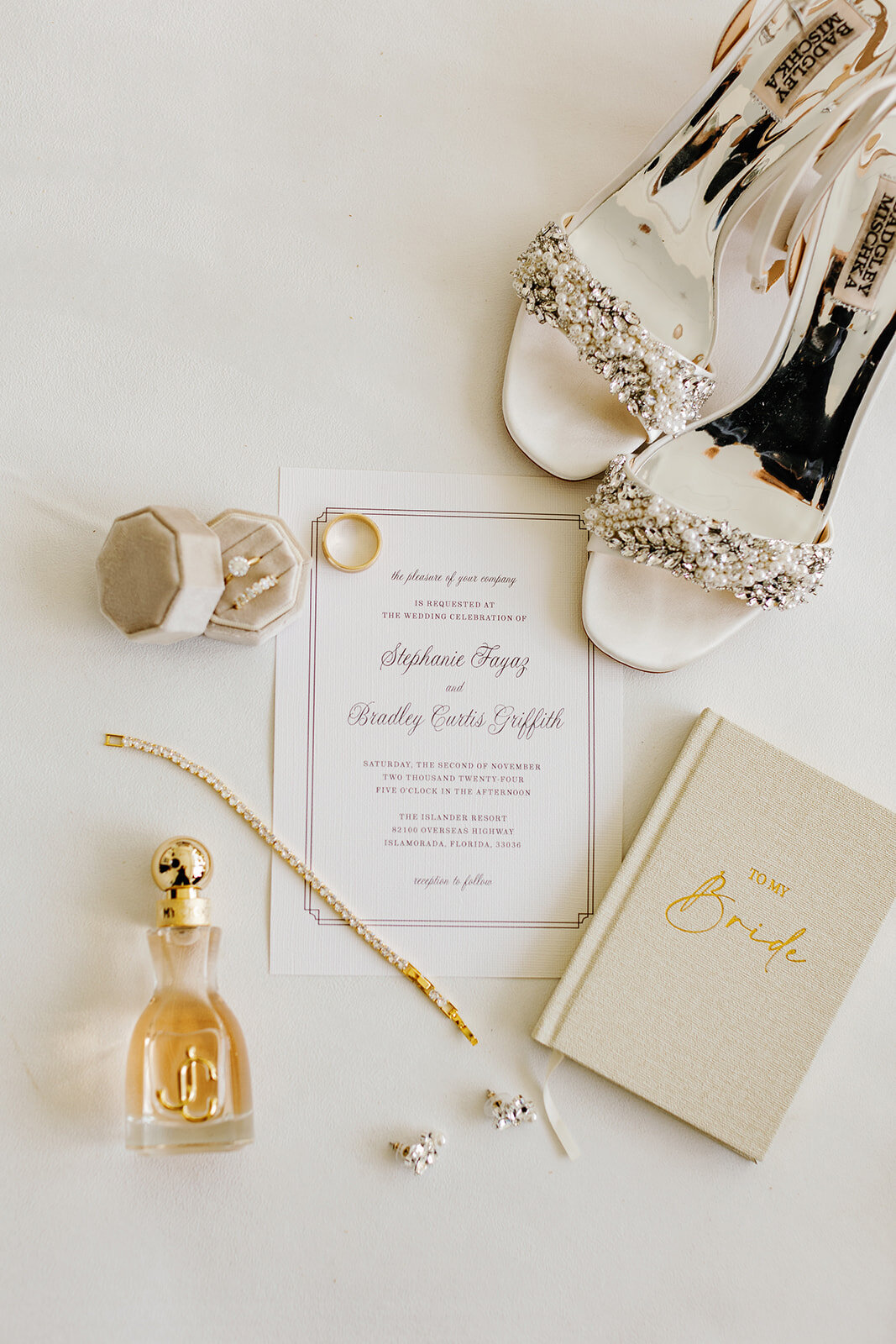 A beautifully styled flat lay featuring Stephanie and Bradley’s wedding invitation, gold wedding bands, a diamond engagement ring, elegant bridal heels, a perfume bottle, a pearl bracelet, and a "To My Bride" vow book. Captured in Islamorada by Claudia Amalia Photography.