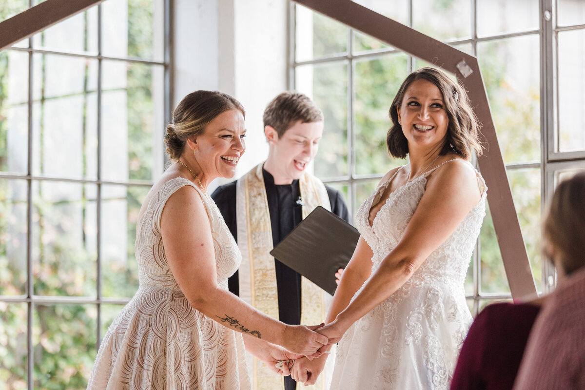 Two brides holding hands and looking out at our their guests during their wedding ceremony at the Hickory Street Annex in Dallas, Texas. Both brides are wearing sleeveless, intricate, white dresses, and their officiant stands behind them while reading from a folder.
