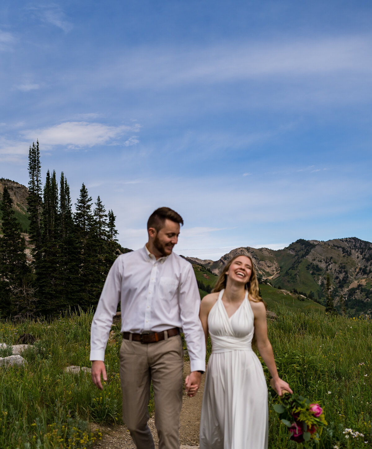 Bride and groom are walking hand in hand towards the camera. The groom is smiling and looking at his bride. The bride is laughing and smiling and slightly throwing her head back. There are mountains and pine trees in the background of this summer Utah elopement.