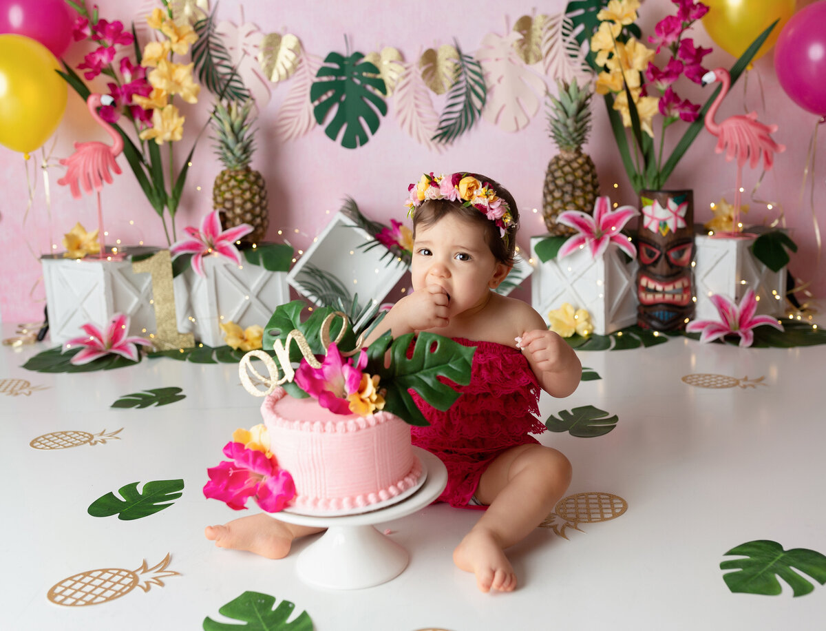 Luau themed cake smash at West Palm Beach Photography studio. Baby girl in red lace romper and floral headband is taking her first bite of cake. The cake is pink and decorated with tropical flowers and leaves. In the background, there is a tropical leaf banner, flowers, pineapples, and flamingos.
