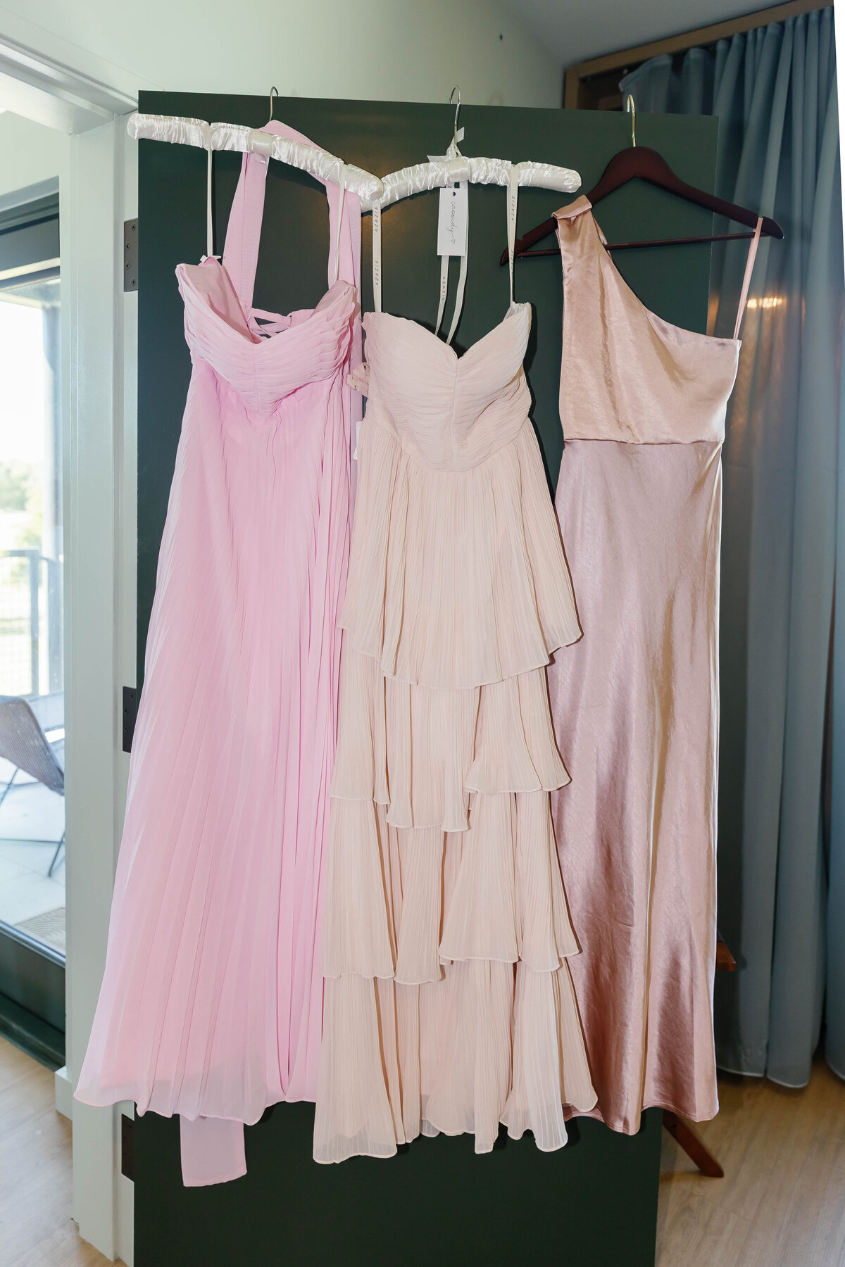 Three bridesmaid dresses in shades of blush pink hanging on a door, styled for a modern elegant wedding, featuring pleated, tiered, and satin fabrics in coordinated pastel tones.
