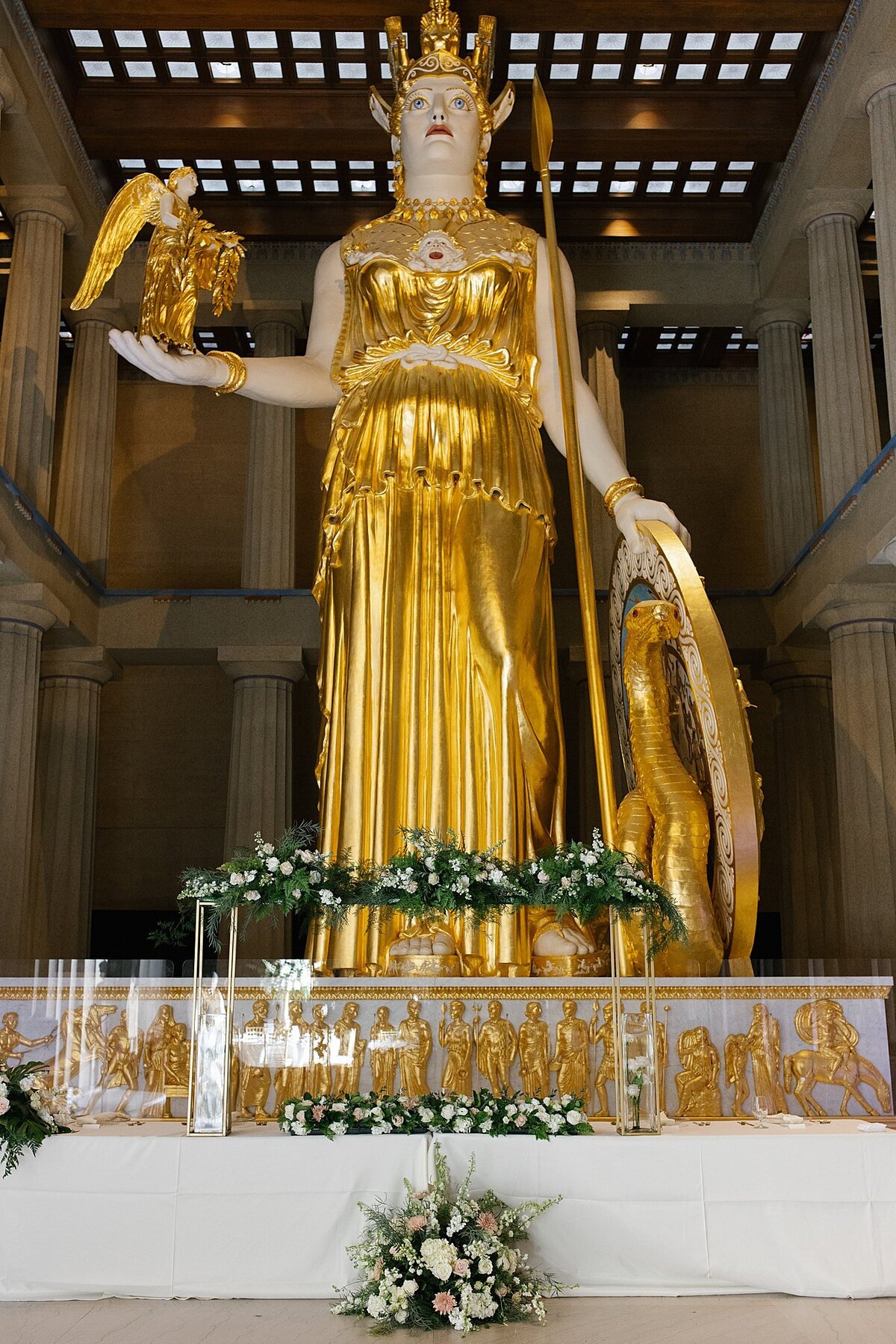 The Athena Statue at The Parthenon in Nashville behind a head table set for a wedding party with white and blush flowers below the table, white and blush flowers on the table and a canopy of flowers above the table on a clear acrylic stand.