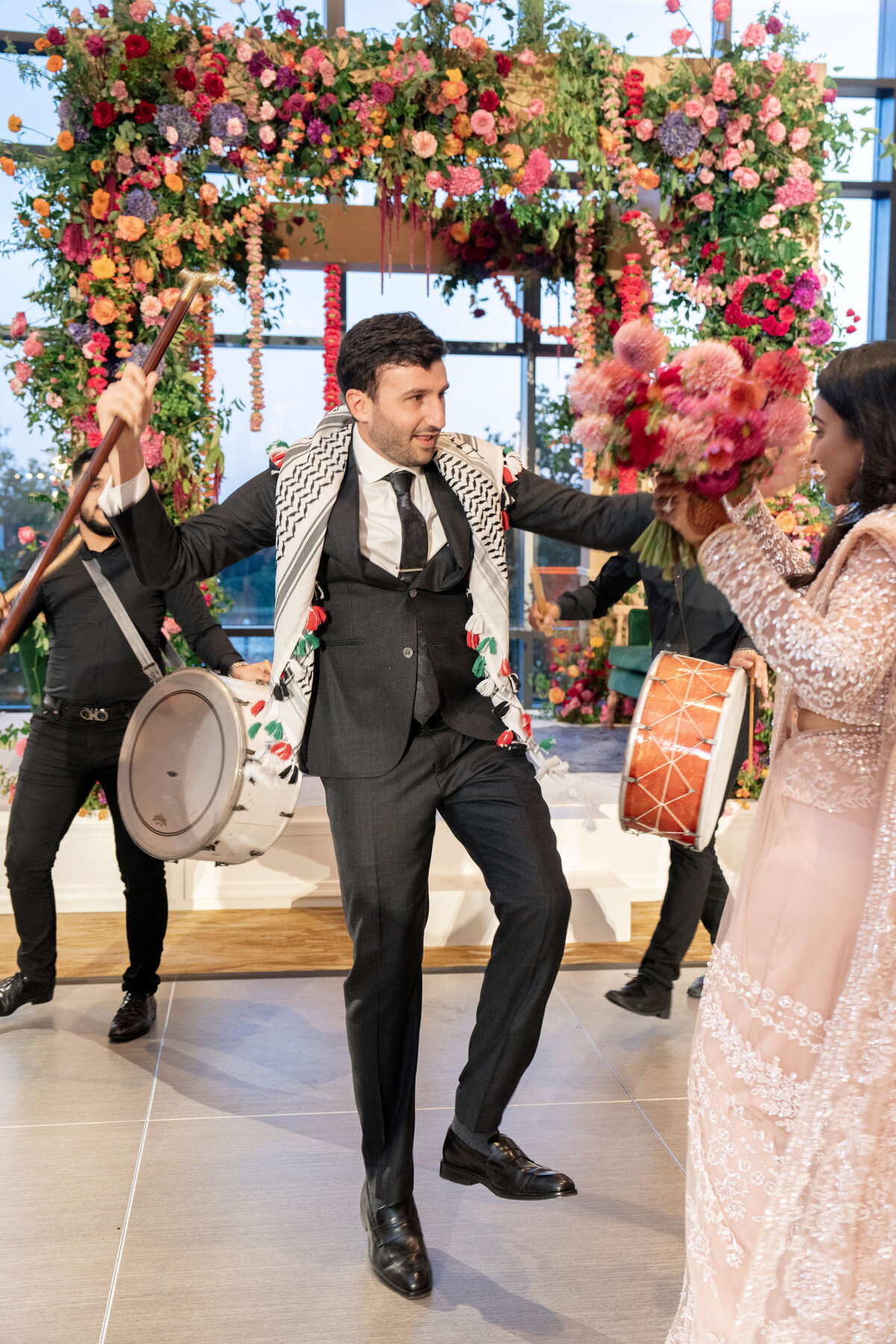 A man in a suit dances joyfully under a colorful floral arch. Hes surrounded by drummers wearing black, and a woman in a light-colored, ornate dress holds a bouquet. The scene is festive and vibrant.