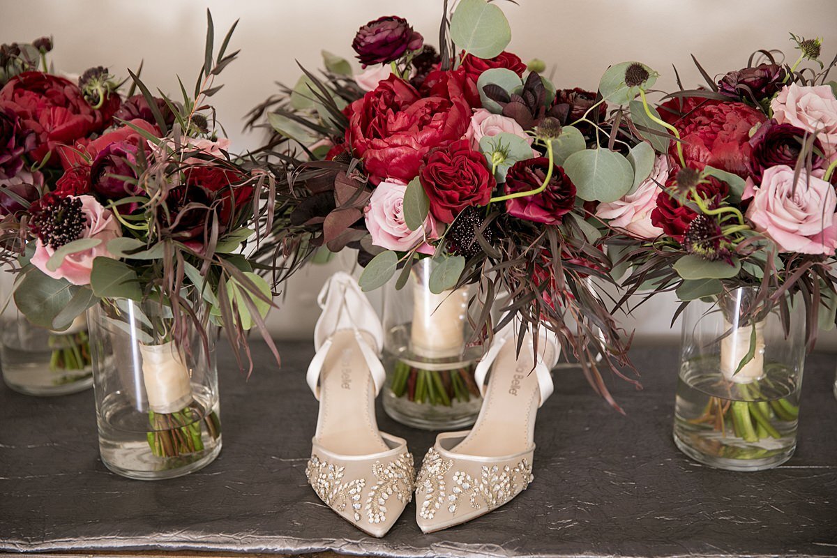 The bride's shoes decorated with delicate vines made from rhinestones on ivory heels placed in front of the bridesmaids bouquets of red peonies, red and blush roses, burgundy flowers with hints of greenery wrapped in an ivory satin ribbon in glass vases at Saddle Woods Farm