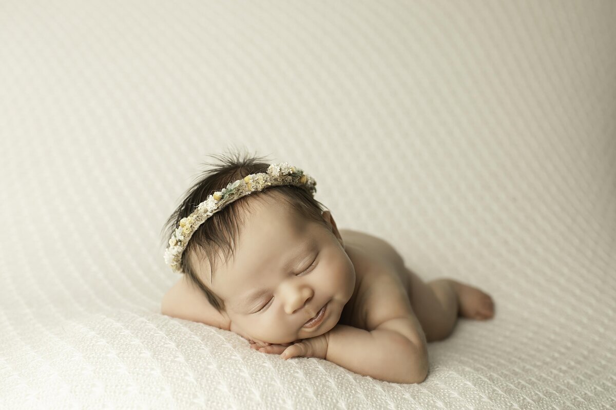 ewborn baby girl resting on a plush white fabric, adorned with a delicate headband. Her tiny hands and soft features add to the sweetness of the moment