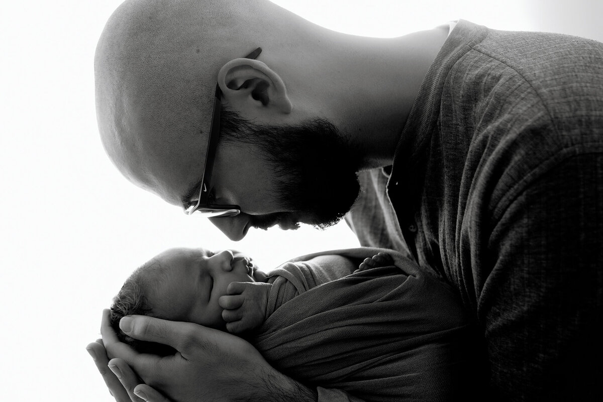 A black and white image from a Pella Newborn Photographer of a dad leaning over his newborn baby son