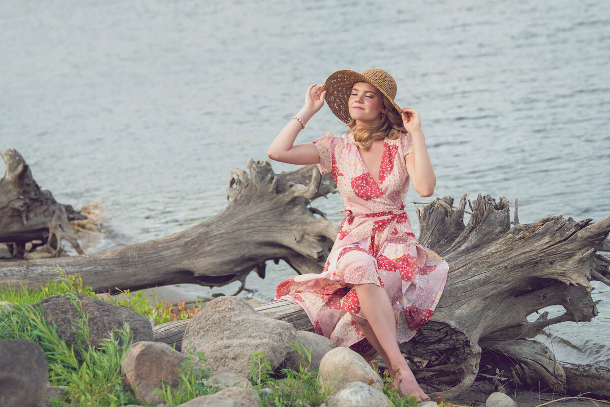 girl in sun hat and flower print dress on driftwood on side of river