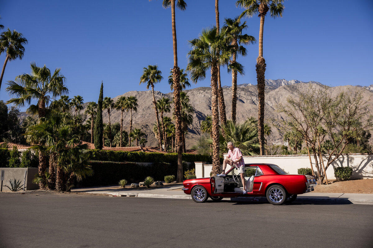 Classic convertible driving through Palm Springs with couple inside, palm trees and mid-century homes lining the sunny street.