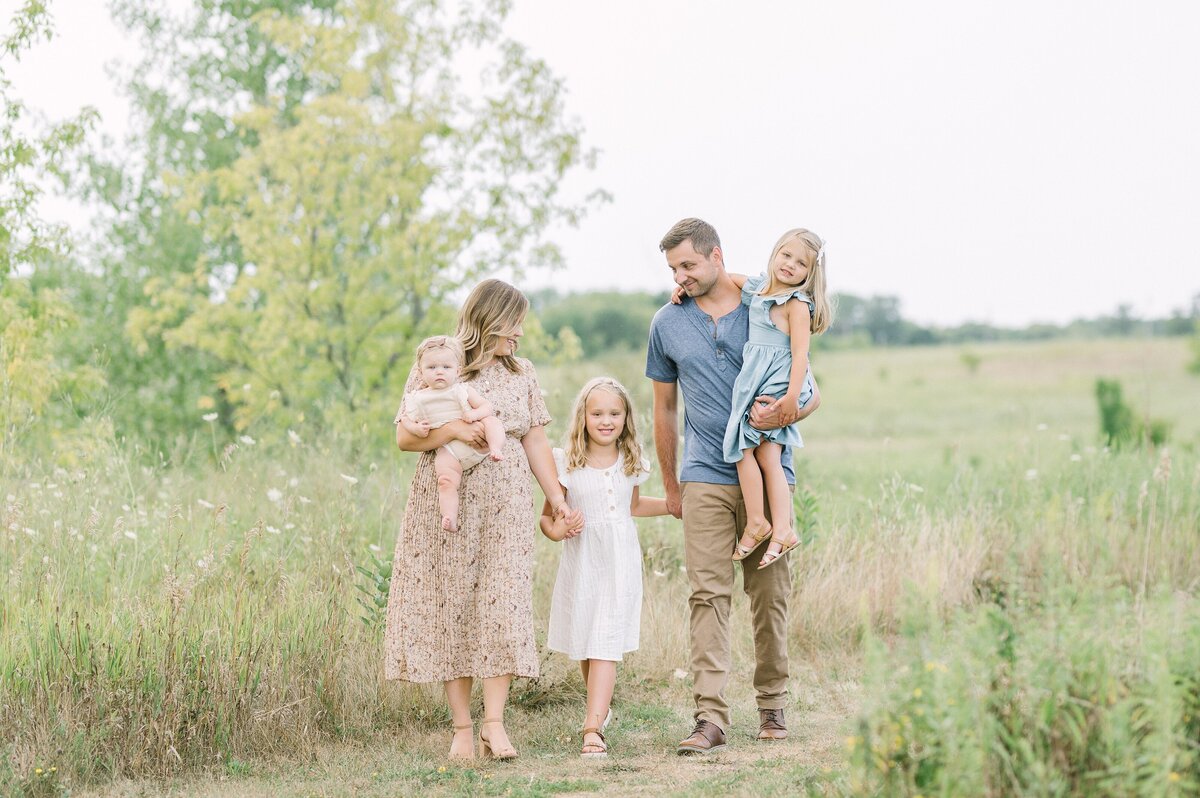 A mom and dad and their 3 girls walk along a grassy path. One of the girls is held by her dad, and one by her mom.