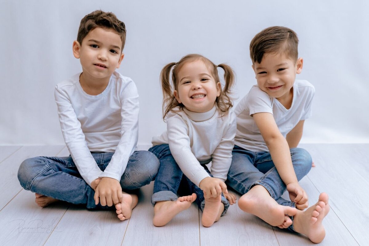 A group portrait of three siblings sitting side-by-side on a light-colored wooden floor. The older brother on the left and the little sister in the middle are looking directly at the camera, while the younger brother on the right is looking to his left, smiling. The two brothers wear white long-sleeved shirts and blue jeans, while the little girl wears a white long-sleeved shirt, blue jeans, and her hair in pigtails.
