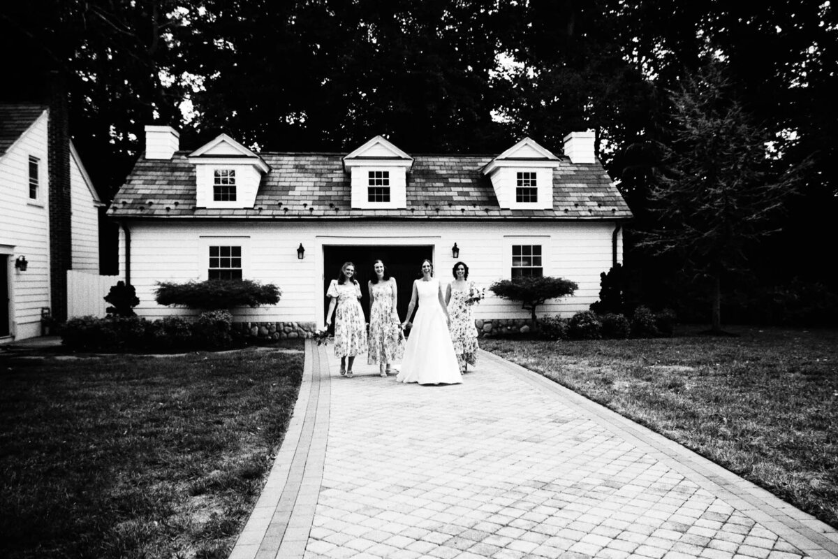 Four women in dresses, including a bride in a white gown, walk down a paved driveway toward the camera in front of a white house with dormer windows, surrounded by trees. Captured by an NJ wedding photographer, the black and white image exudes timeless elegance.