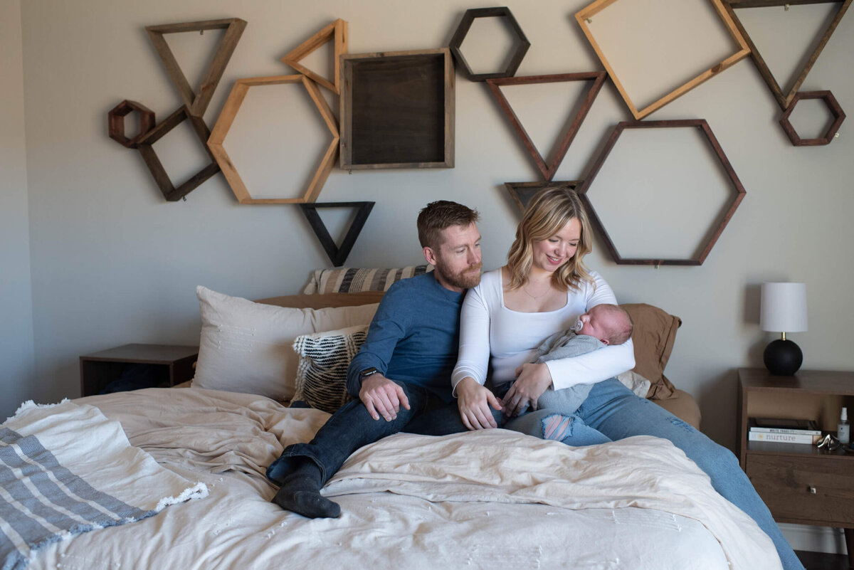 New mom and dad sit on a bed smiling at their newborn baby in mom's lap