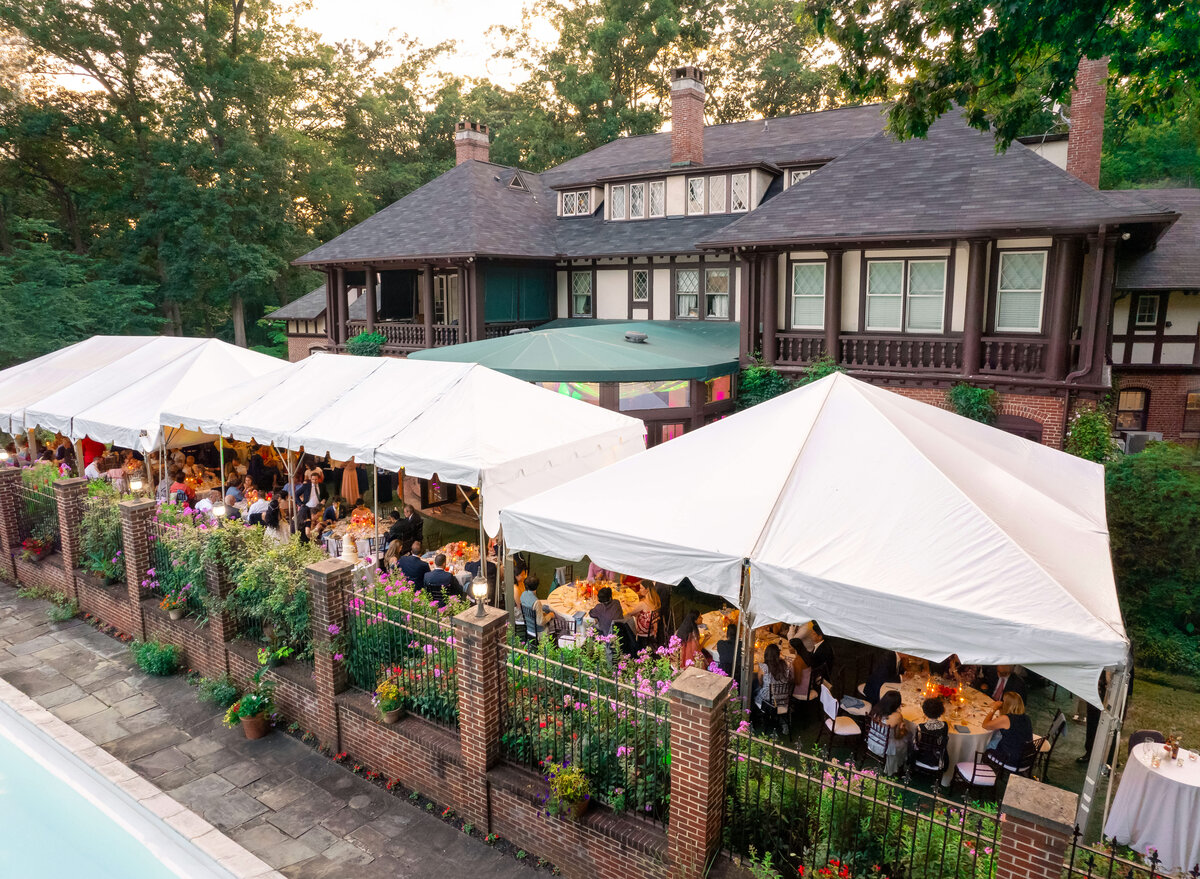 A large outdoor event is taking place under white tents beside a grand house. Guests are seated at tables, surrounded by lush greenery and flowers. The setting includes a brick patio and a nearby swimming pool, with trees in the background.