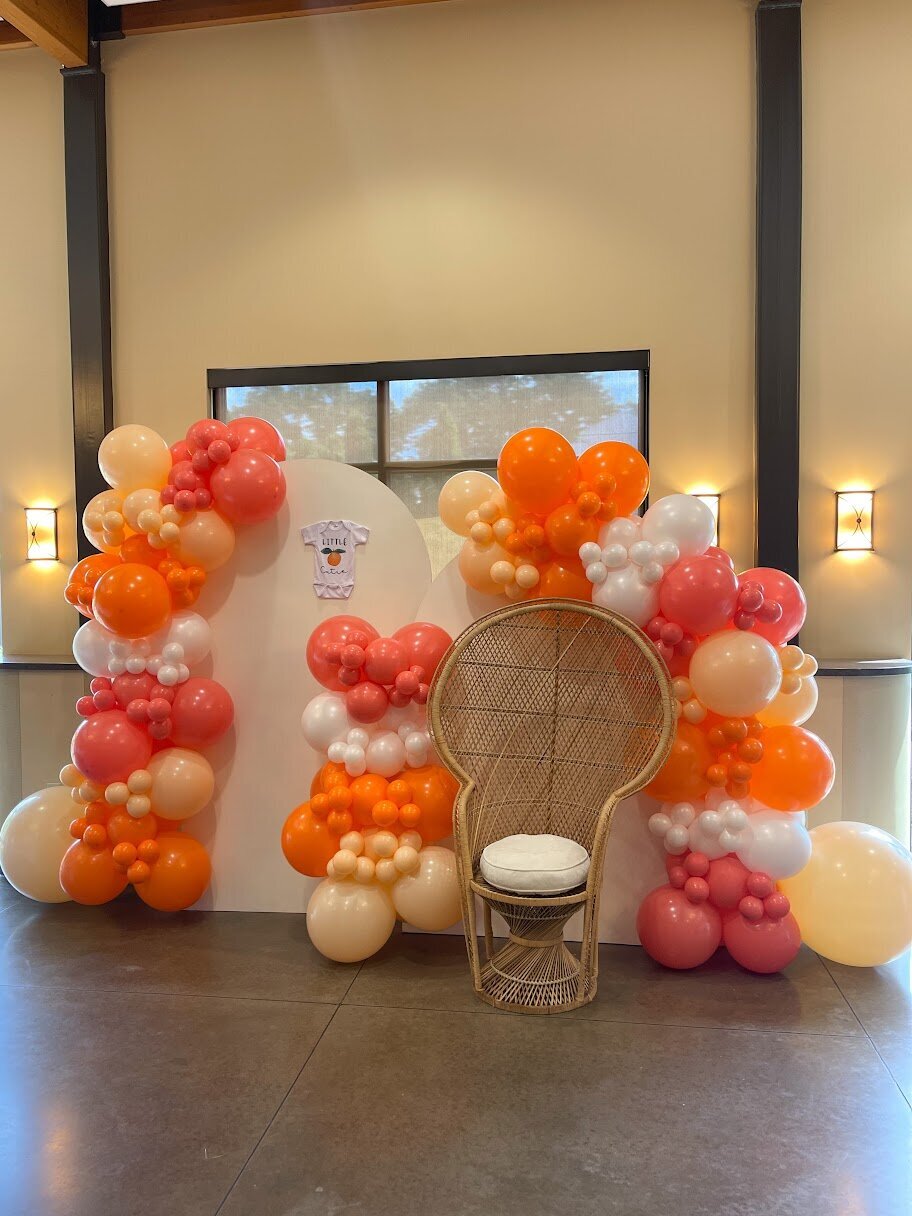 Orange and white balloons set up around white wooden arches and a peacock chair.