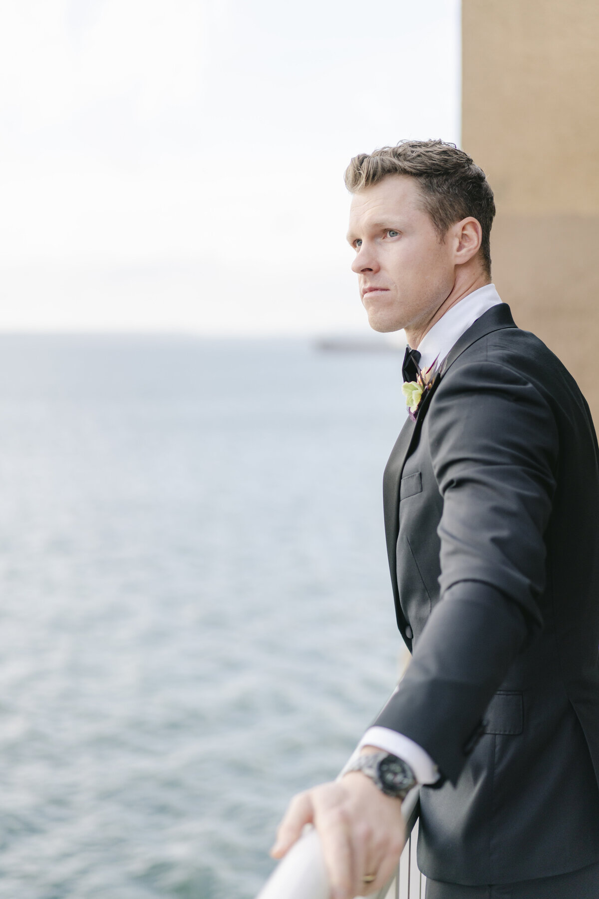 a groom holding a railing and looking off into the distance at the ocean in california