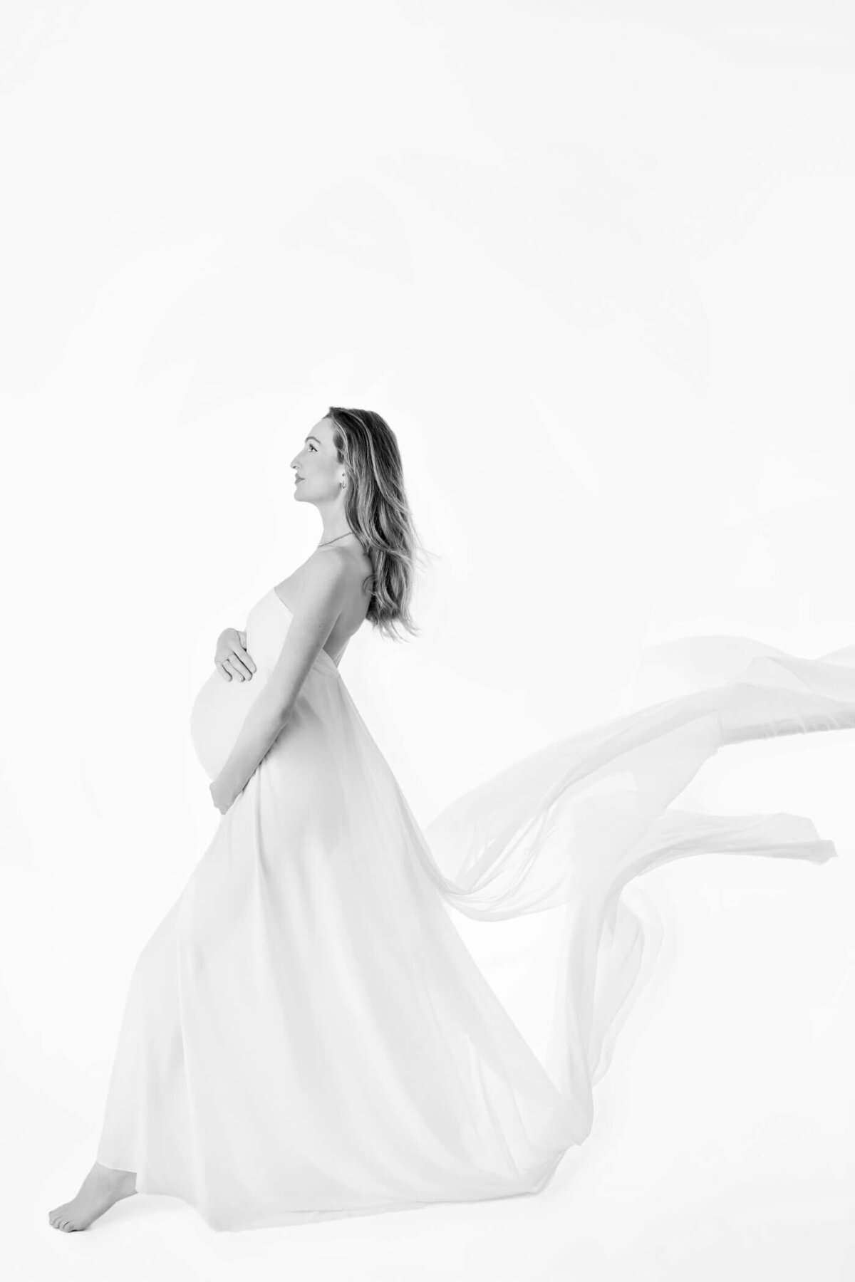 A black-and-white profile image of a pregnant woman standing barefoot against a stark white background. She wears a flowing, sheer gown that wraps around her and billows dramatically behind her, as if caught in a gentle breeze. Her hands rest on her baby bump, one above and one below, as she gazes forward with a serene expression. The image captures the ethereal beauty and grace of pregnancy, emphasizing the flowing lines of the gown and the calmness of the moment.