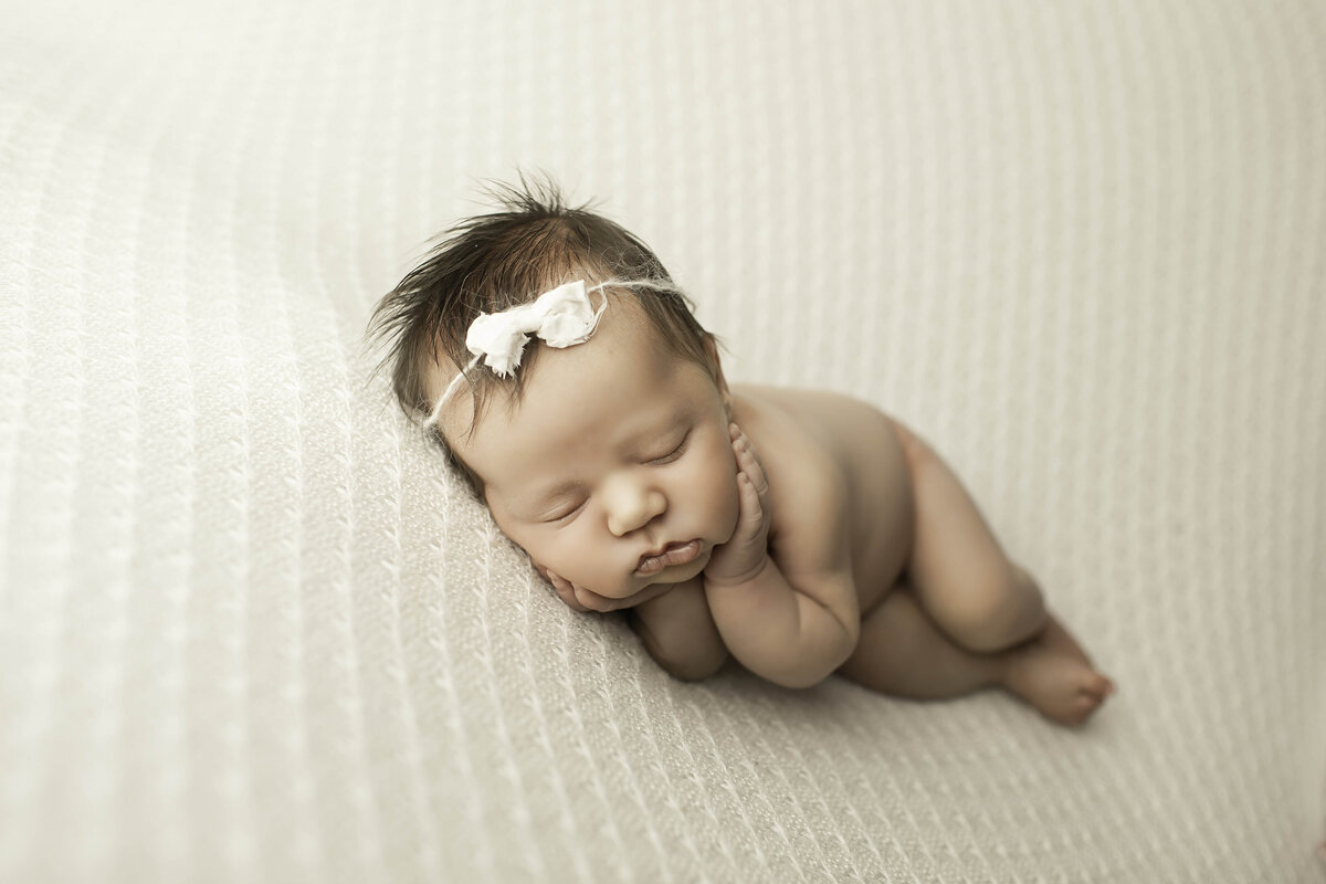 Newborn baby girl peacefully lying on soft white fabric, wearing a dainty headband. Her tiny features and serene expression create a delicate and timeless look
