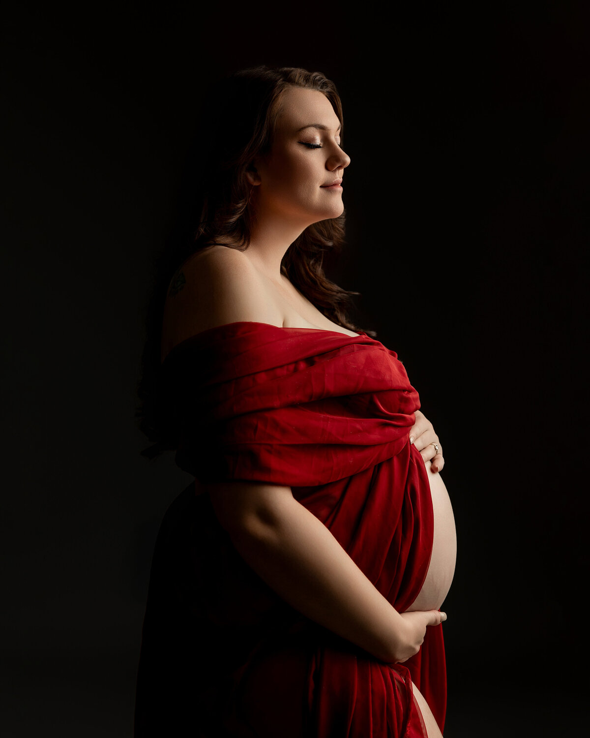 A radiant expectant mother dressed in a bold red gown, captured in a close-up shot with a dark, moody backdrop. Captured by Inland Empire maternity photographer Kaitlyn Dawn Photography.