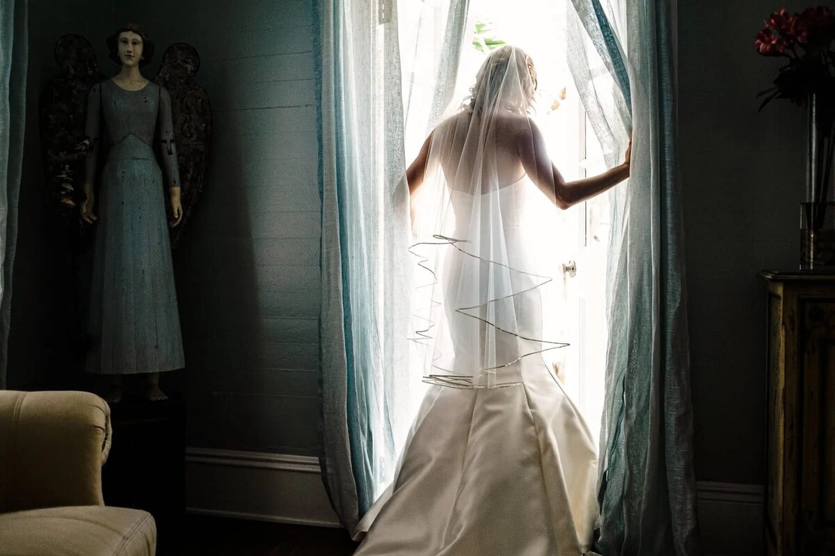 A bride in a white gown stands at a window, backlit by soft light, with an angel statue in the background