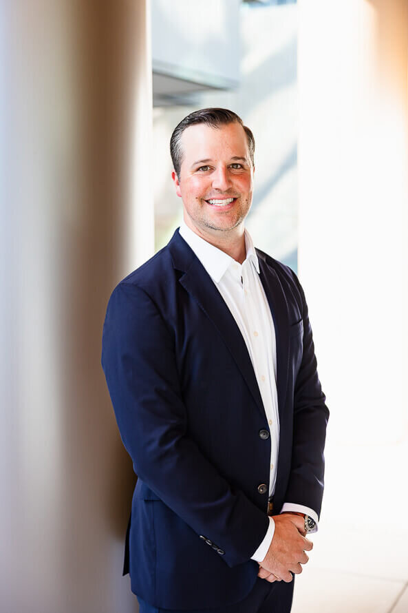 Man in dark blue coat folds hands and smiles for his headshot outside.