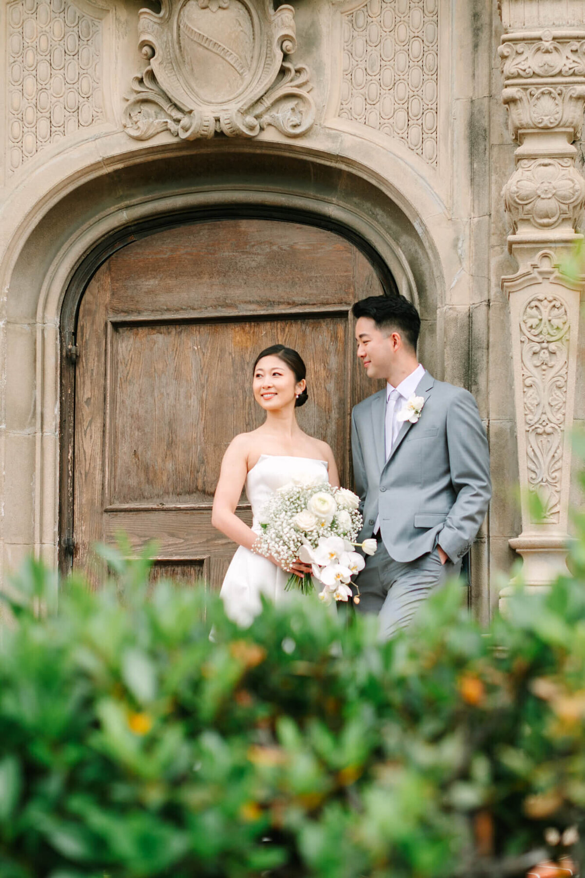 Bride in white dress holding a bouquet stands beside groom in a gray suit. They smile at each other in front of an ornate wooden door, surrounded by greenery.
