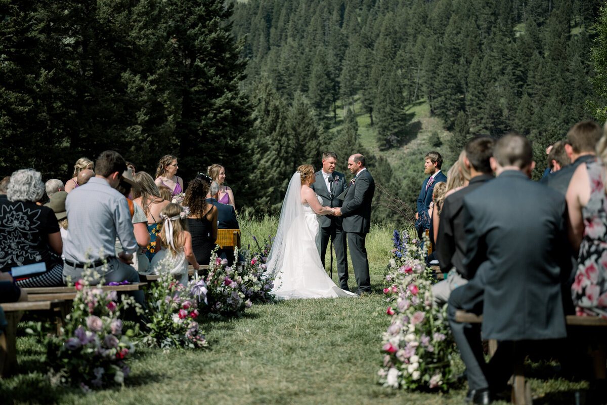 Bride and groom sharing vows surrounded by family and friends with the Montana mountains as a backdrop during their summer wedding.