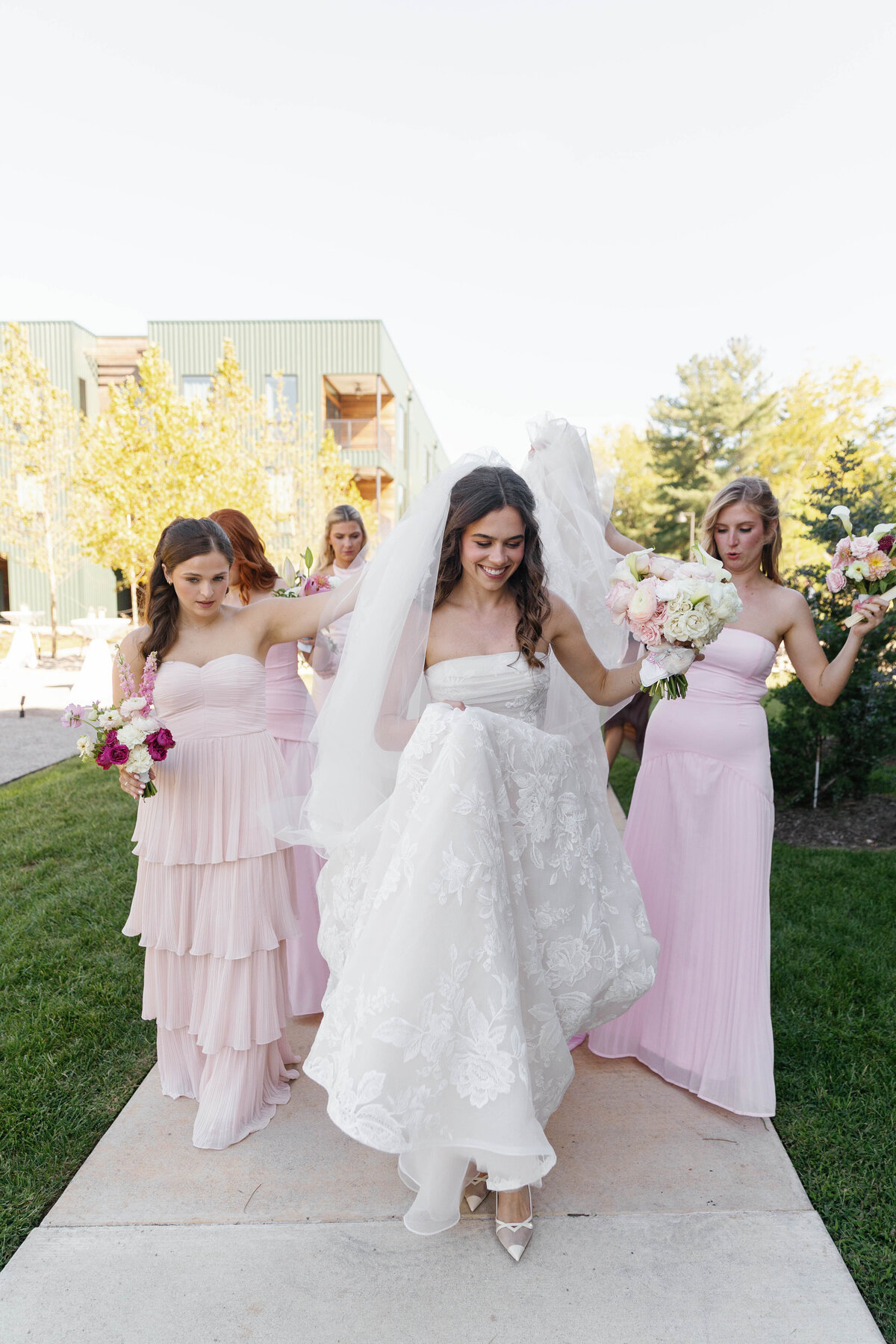 Bride walking outdoors with her bridesmaids in soft pink dresses, holding her lace wedding gown and bouquet during a spring wedding at a modern venue, natural light, joyful candid moment.