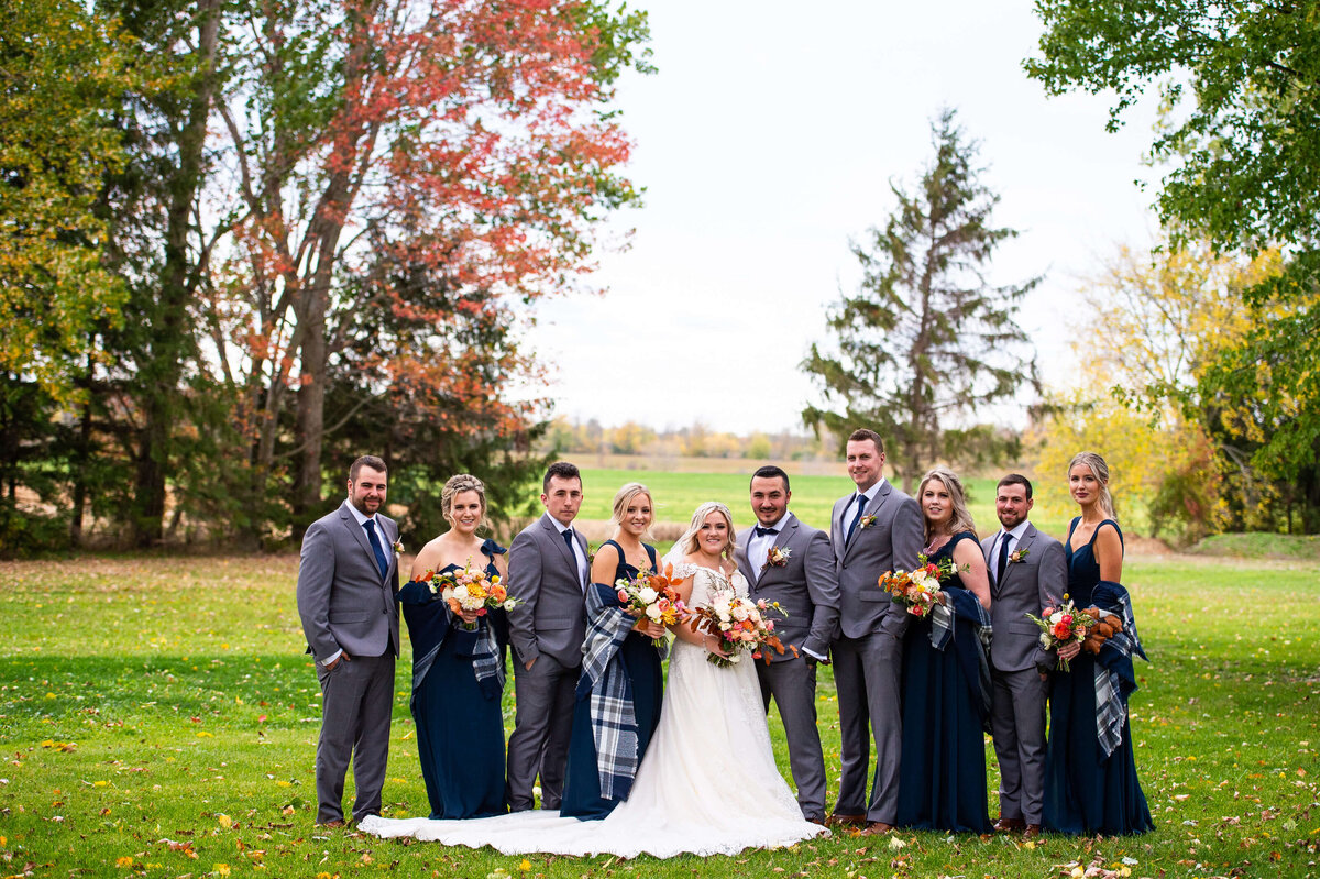 an Ottawa wedding bridal party with the bridesmaids wearing  long navy gowns holding fall bouquets and the groomsmen in light grey suits.  Taken outside at Strathmere wedding venue in Ottawa