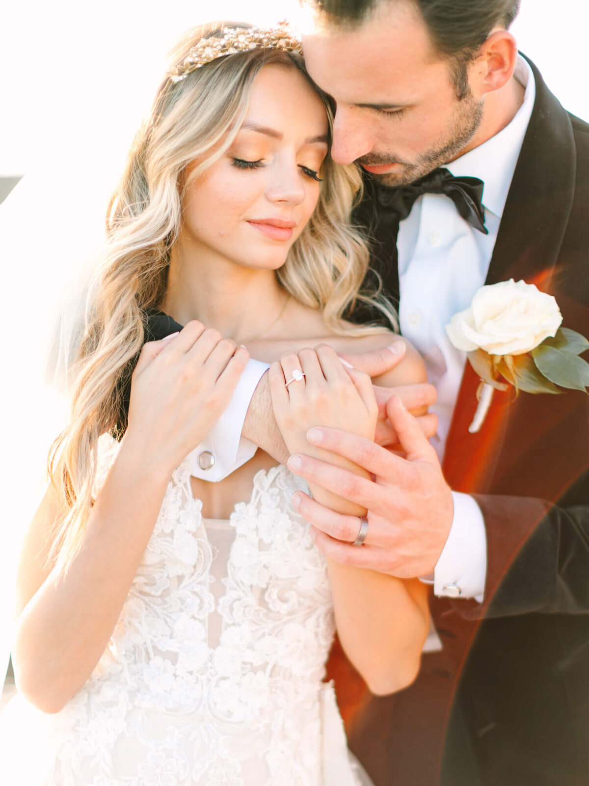Bride in a lace gown and tiara stands with groom in a black suit holding her hands.