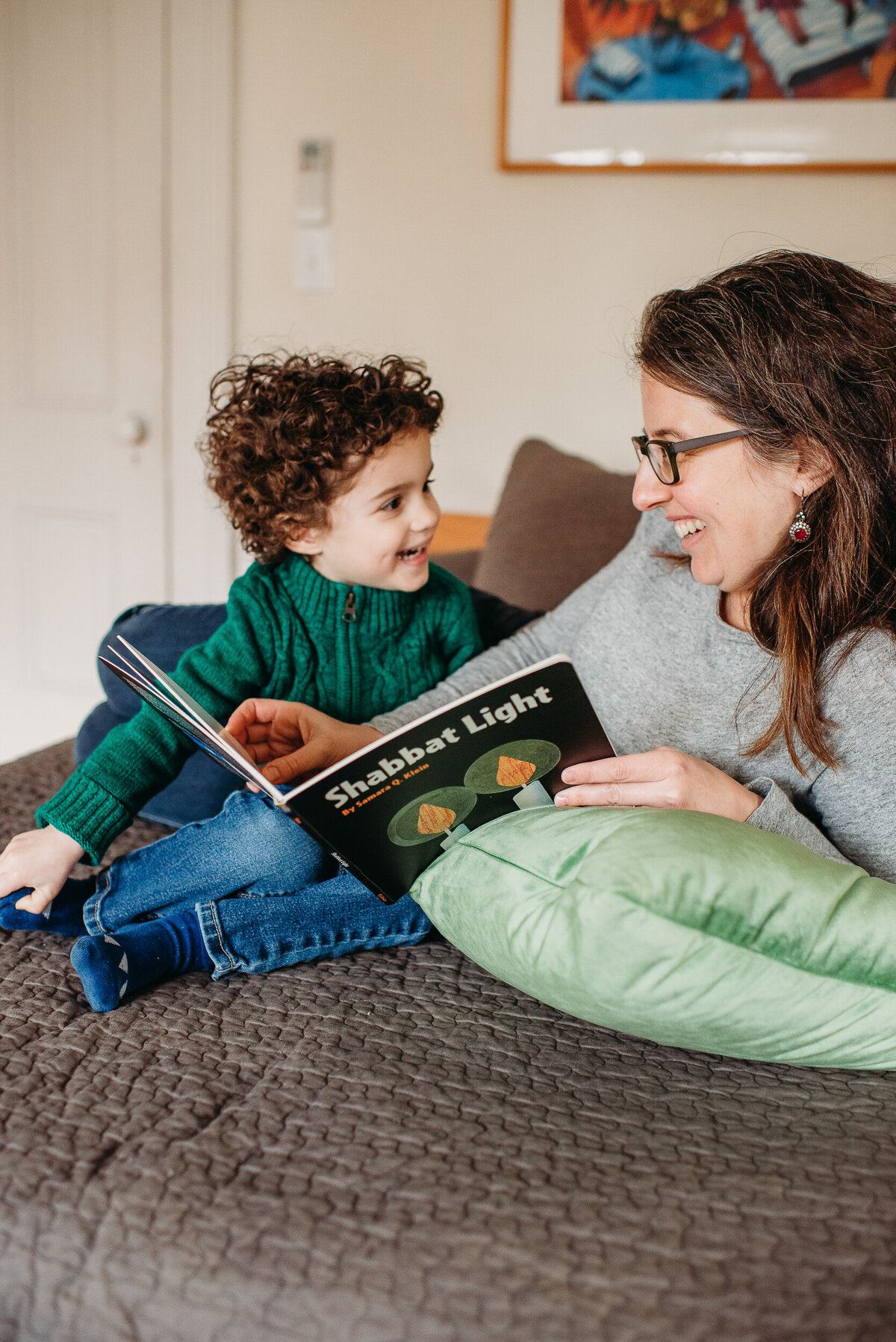 mom smiles at preschool boy while reading story on bed