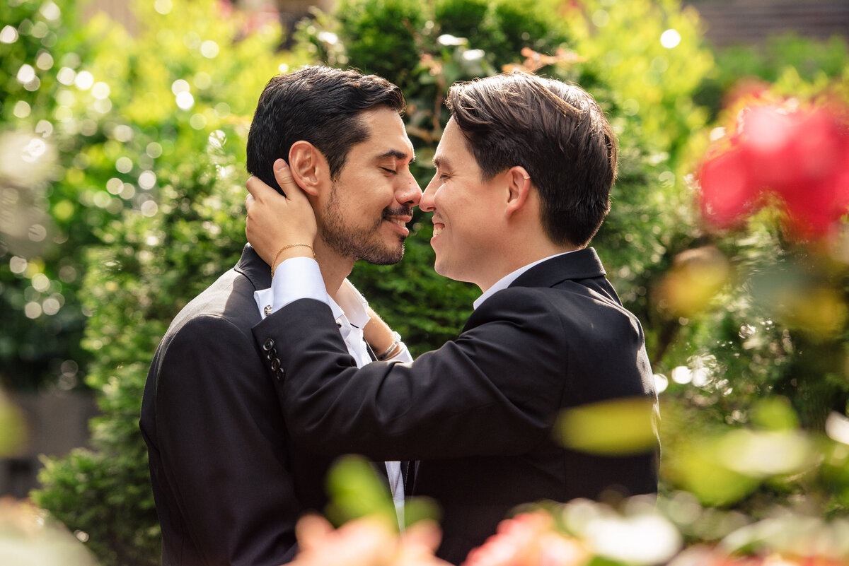 Portrait through foreground leaves  of gay male couple smiling and touching noses in the rose garden at The Duke Mansion by Charlotte wedding photographer DeLong Photography