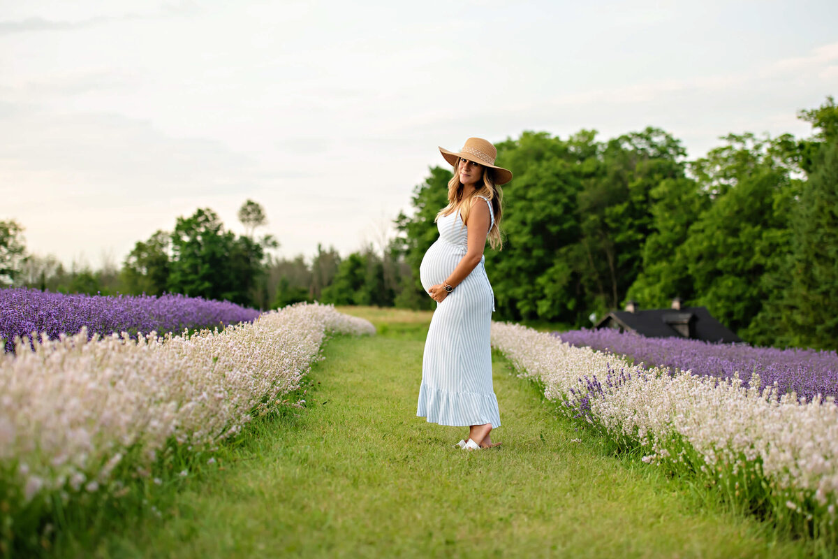 pregnant woman standing in a lavender field wearing a blue and white stripped dress and beige had for her outdoor maternity photos in Burlington