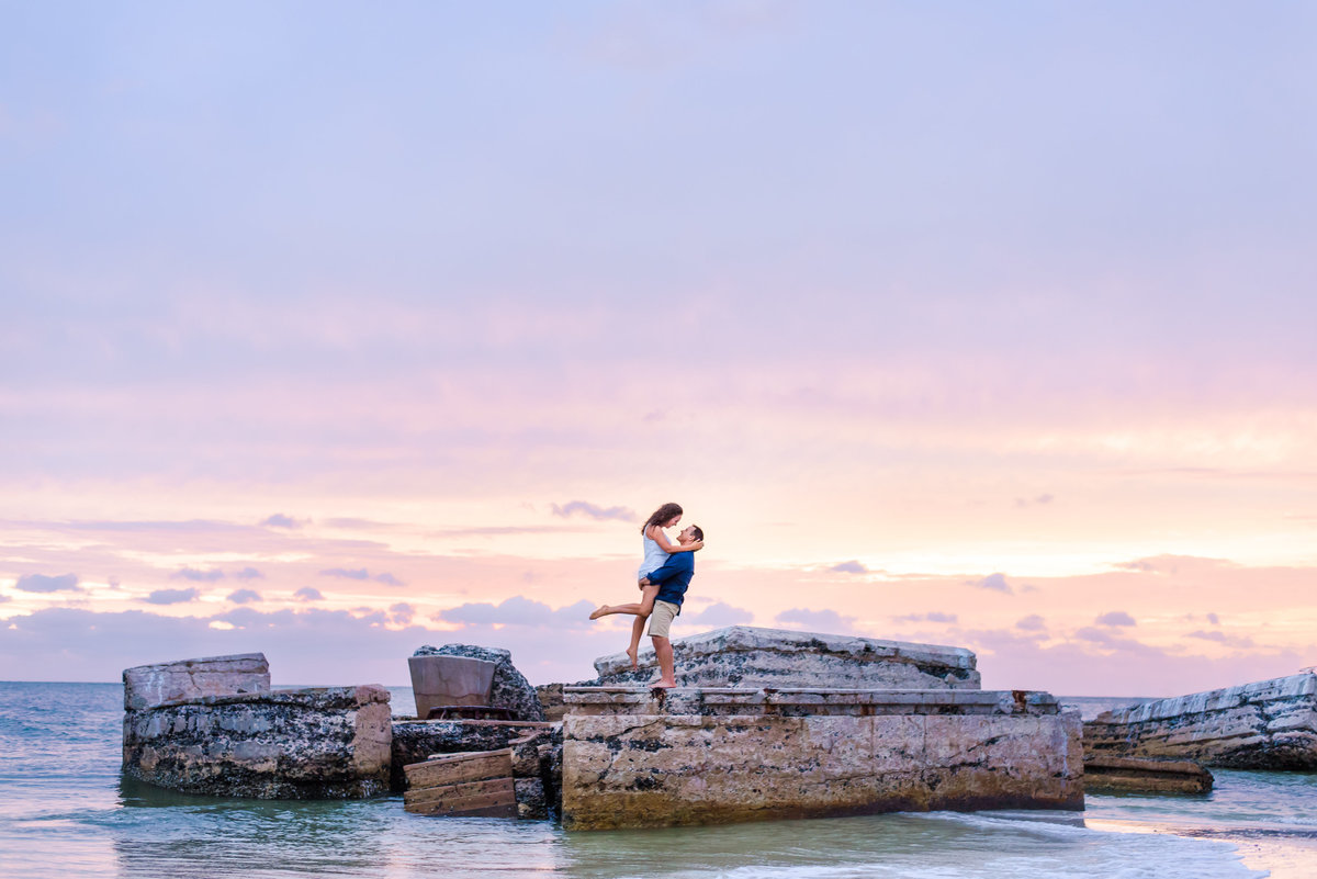 Engaged couple share a romantic hug on a big rock in front of a sunset at Fort Desoto National Monument