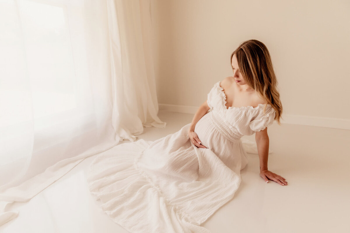 Woman wearing a white long dress with a sweetheart neckline sitting on a white floor while holding her baby bump.