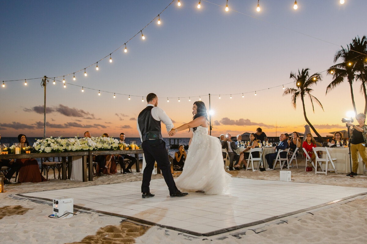 A couple shares their first dance at a beachside destination wedding reception under a string of lights at sunset.
