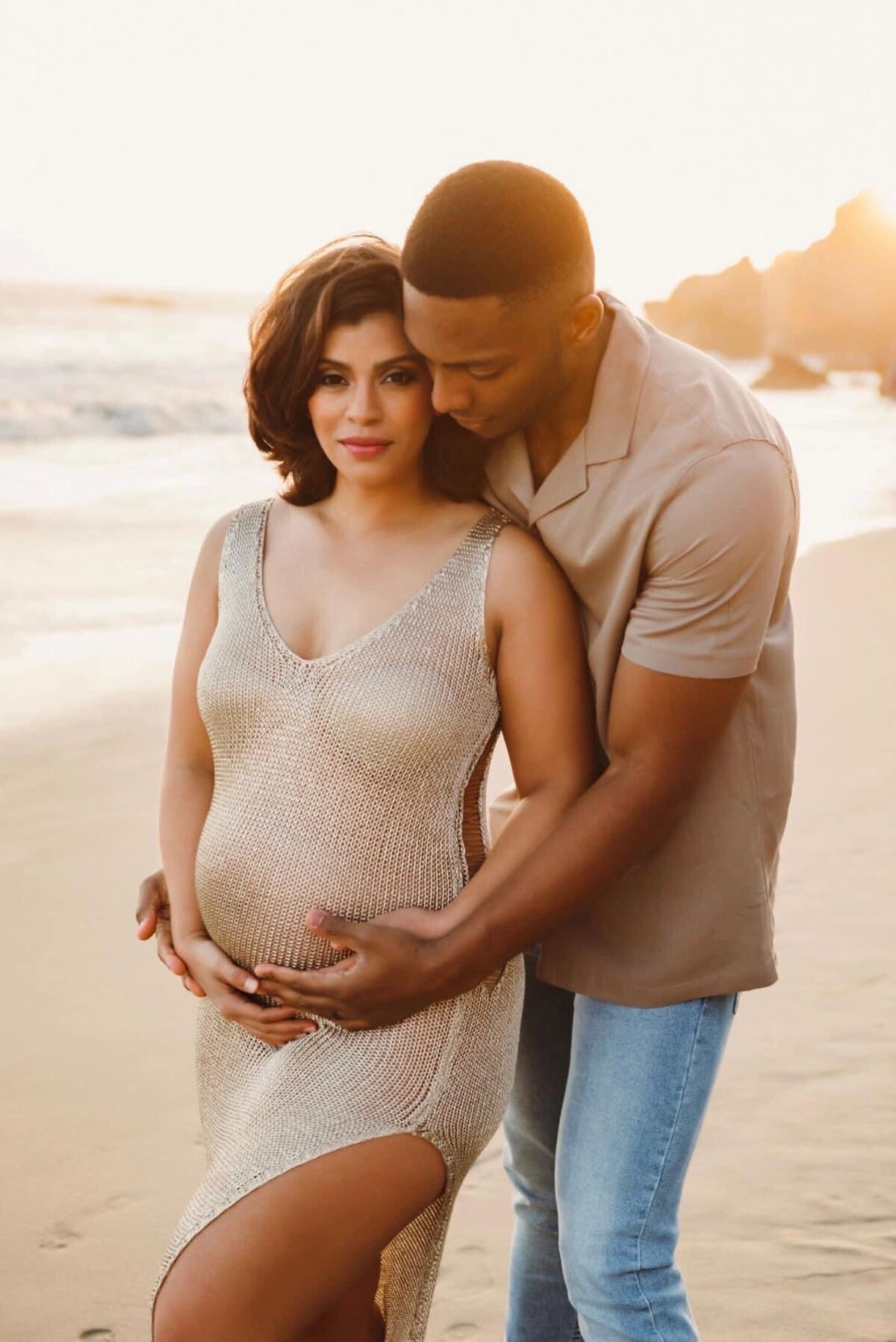 A man and pregnant wife  holding each other on the beach.