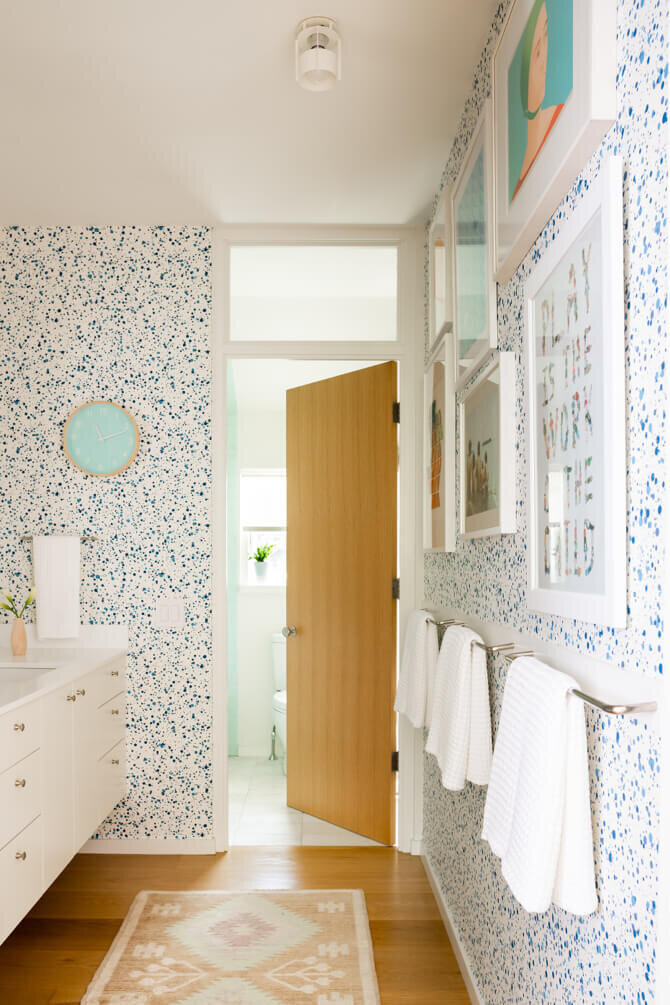 Interior designer in Hingham creates a modern and playful bath with a white vanity, Hinson splatter wallpaper, colorful art, a natural oak door, and blue accents.