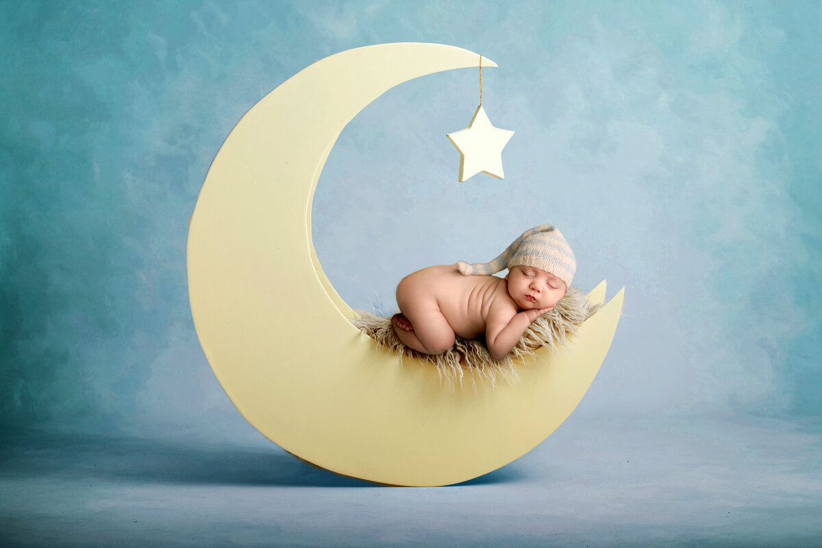 Wide shot of infant asleep on a moon prop in a striped stocking hat.