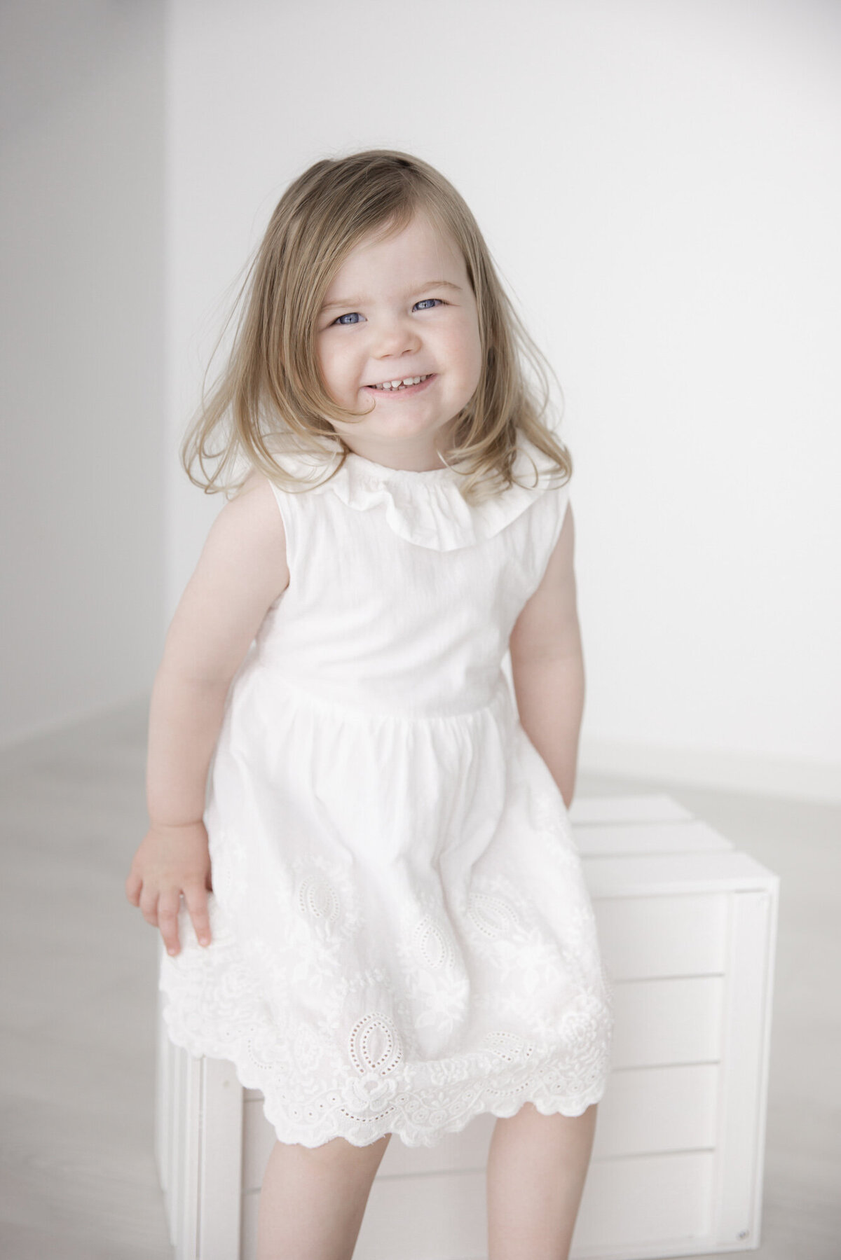 En jente på rundt tre år sitter på en hvit kasse i Studio Landmark og smiler mot kamera. Hun har på seg hvit kjole. Fotografert i Studio Landmark, Fyllingsdalen.