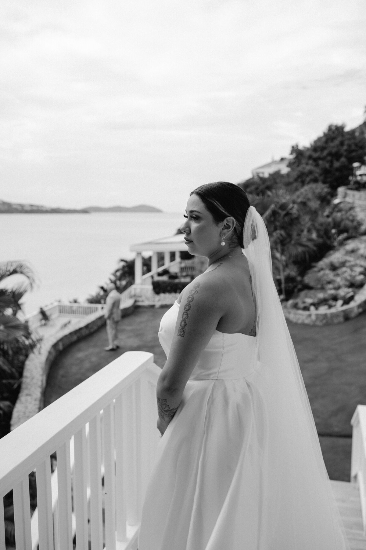 A bride in a white gown looking pensively over a balcony with a scenic ocean view in the background