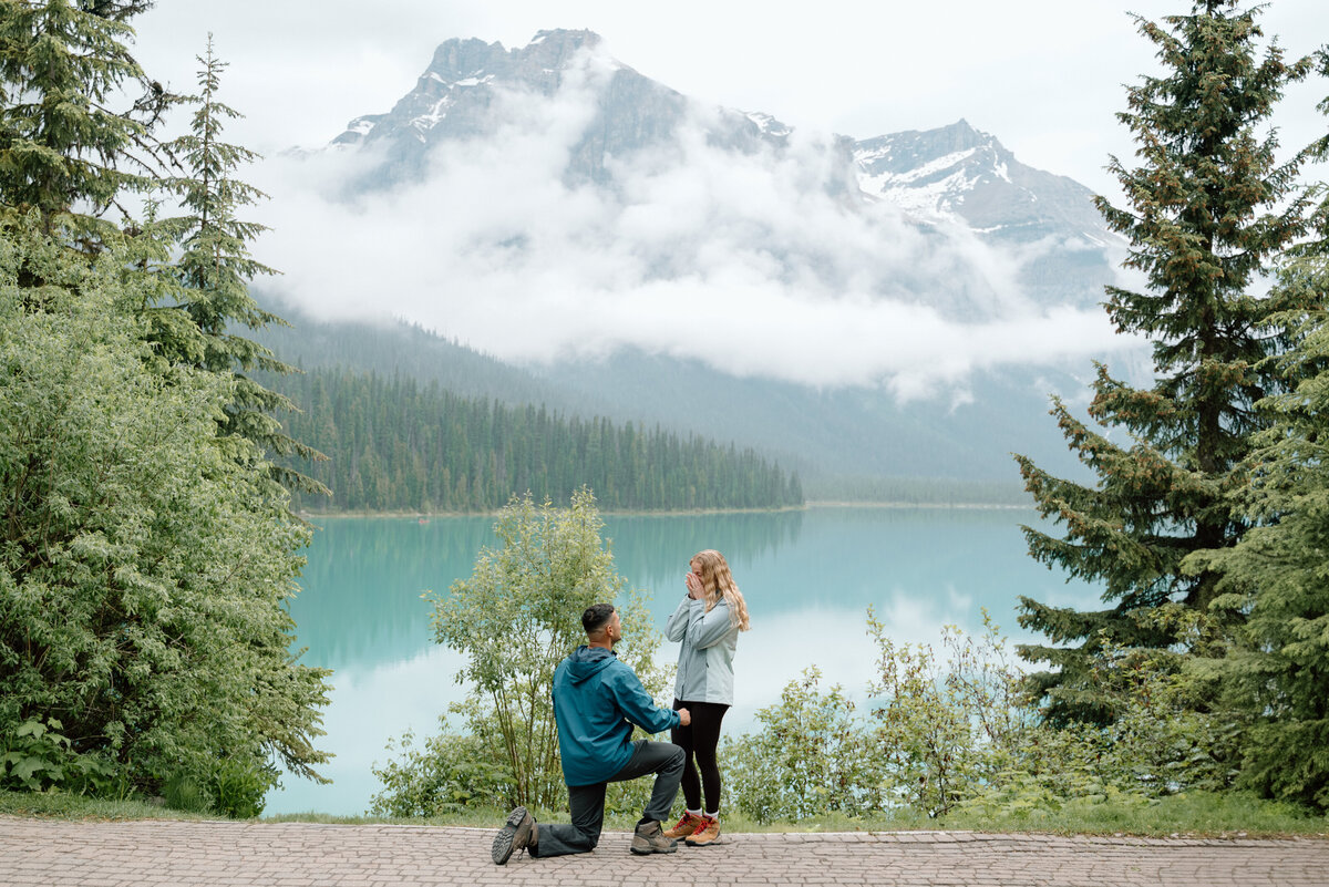 Romantic proposal at Emerald Lake with groom-to-be kneeling in front of turquoise lake and mountains.