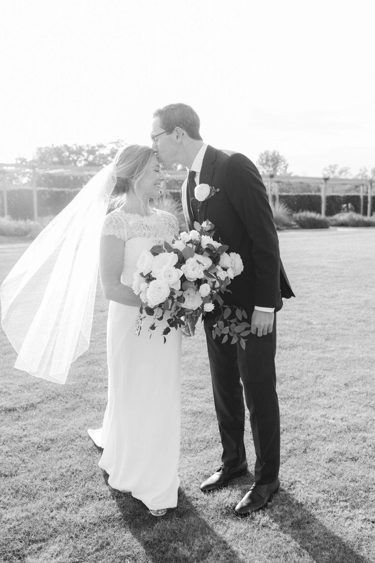 Black and white outdoor photo of groom kissing bride's forehead