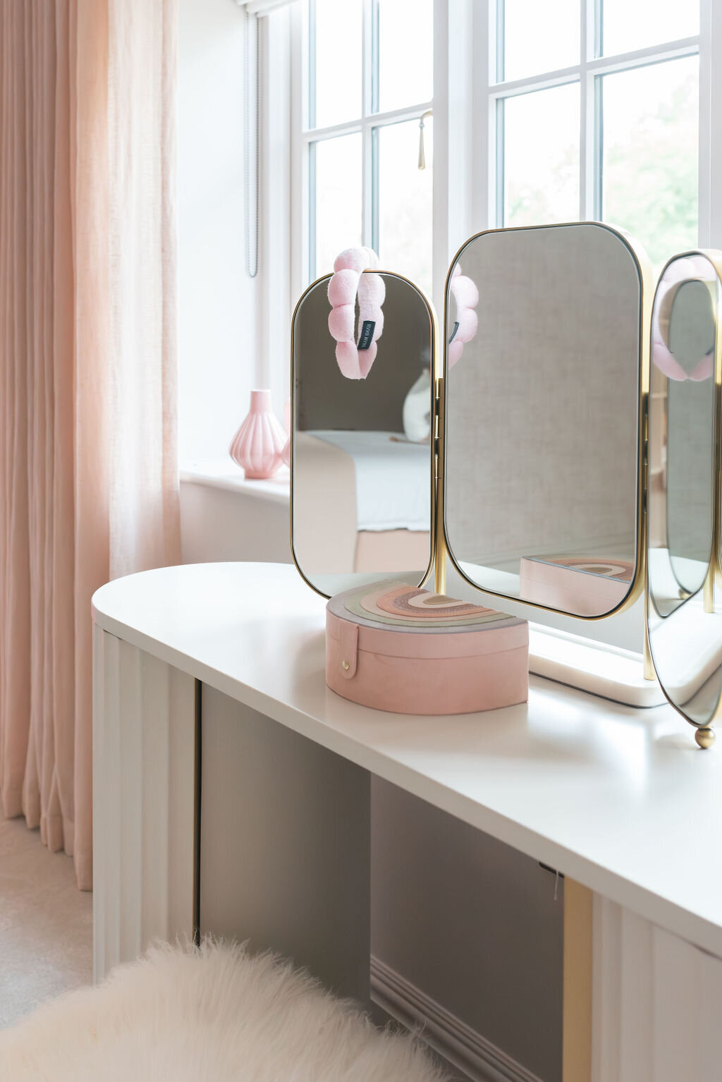 A white vanity table with a tri-fold mirror and a soft pink stool in a girl's pink bedroom.