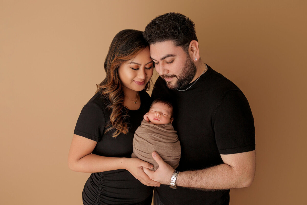 A young couple lovingly holding their swaddled newborn baby, all wearing black clothing against a warm beige background.