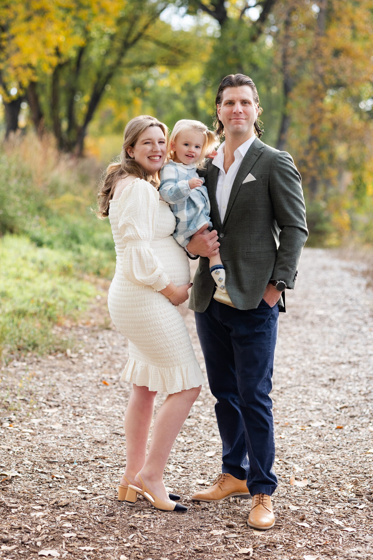 Mom and dad hold their toddler daughter between them and all three smile at the camera as mom holds her pregnant belly with one hand.