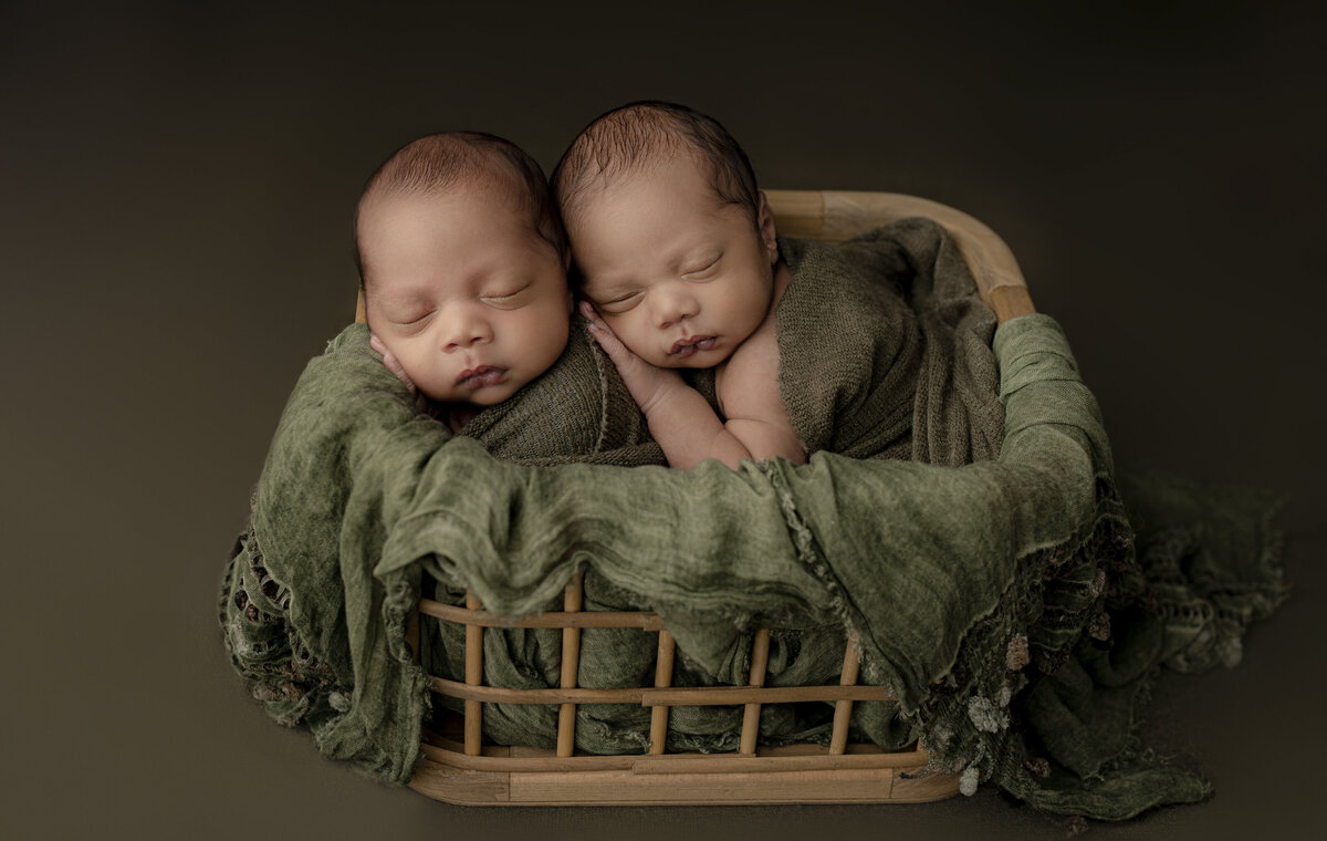 newborn twin boys wrapped up and posed in basket sleeping on each other on green blanket