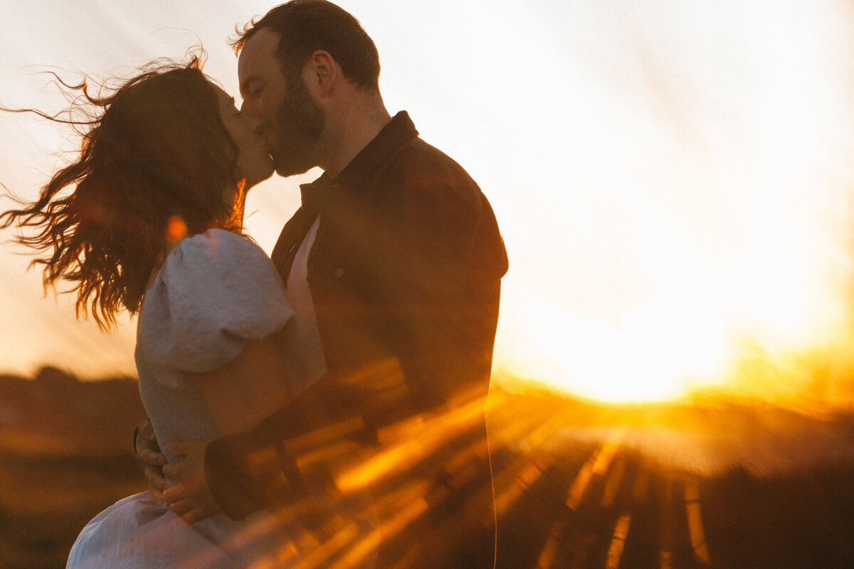 Shot through the dunes at sunset a couple kiss backlit by the golden sunlight