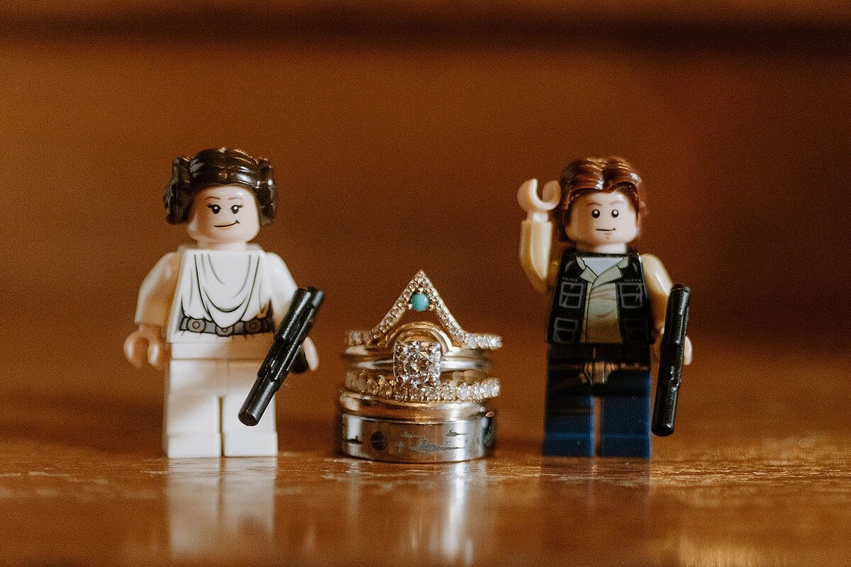 Lego Princess Leia and Lego Han Solo stand on either side of a stack of wedding bands with an engagement ring. The groom's ring is platinum. The bride's wedding ring is a thin gold band. The engagement ring is a set of rings. A diamond infinity band, a round cut diamond solitaire and a triangular shaped ring with diamonds and a small turquoise stone.