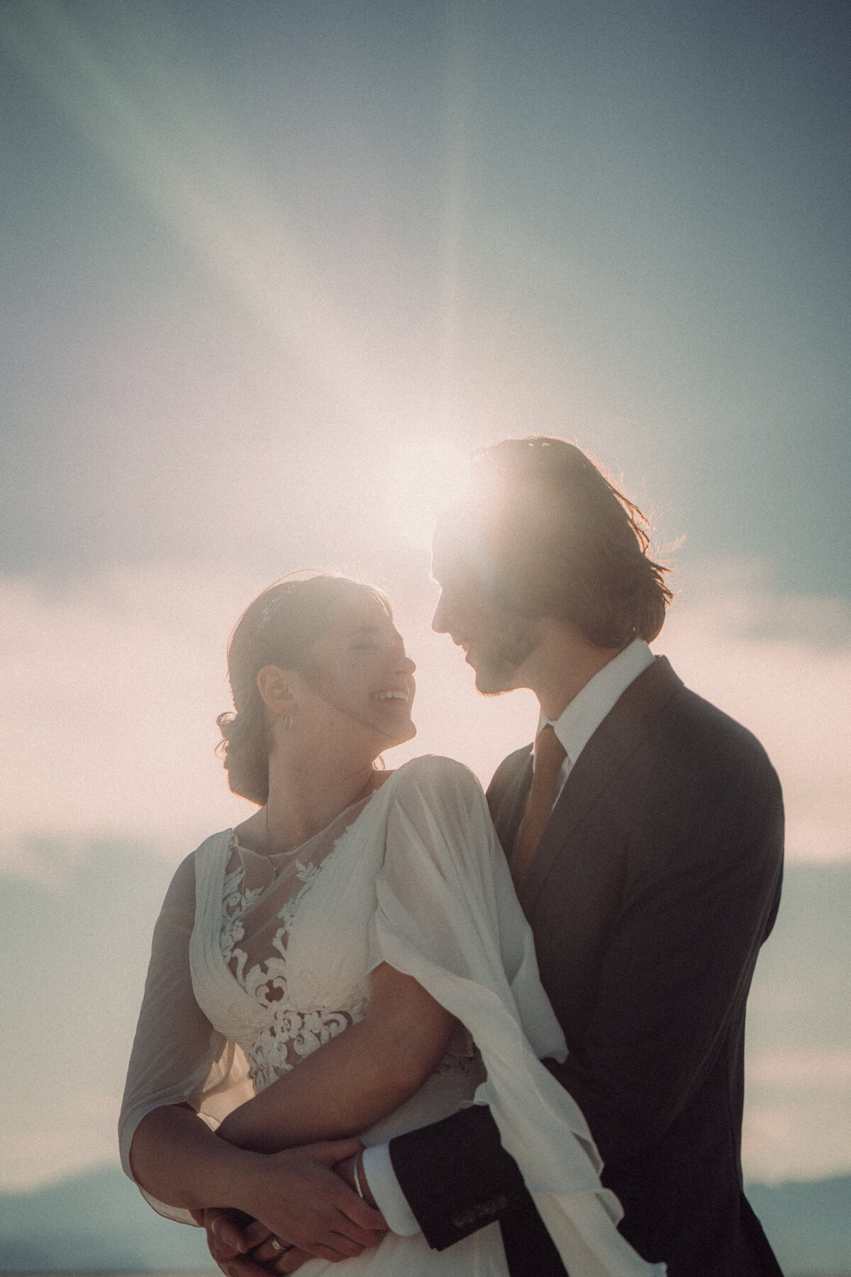 Bride and Groom stand in front of sunset and hold onto each other with the Salt Flats in the background.