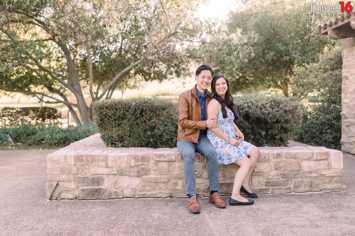 Engaged couple sit together on a stone planter wall